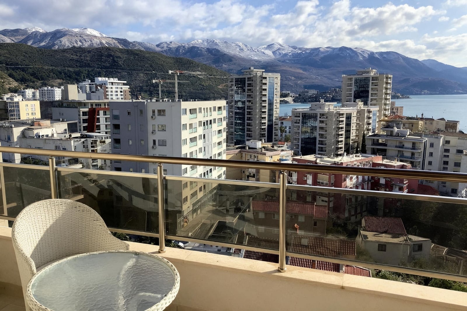 View from a balcony overlooking a city, sea, and snow-capped mountains. Wicker furniture and glass railing are visible.