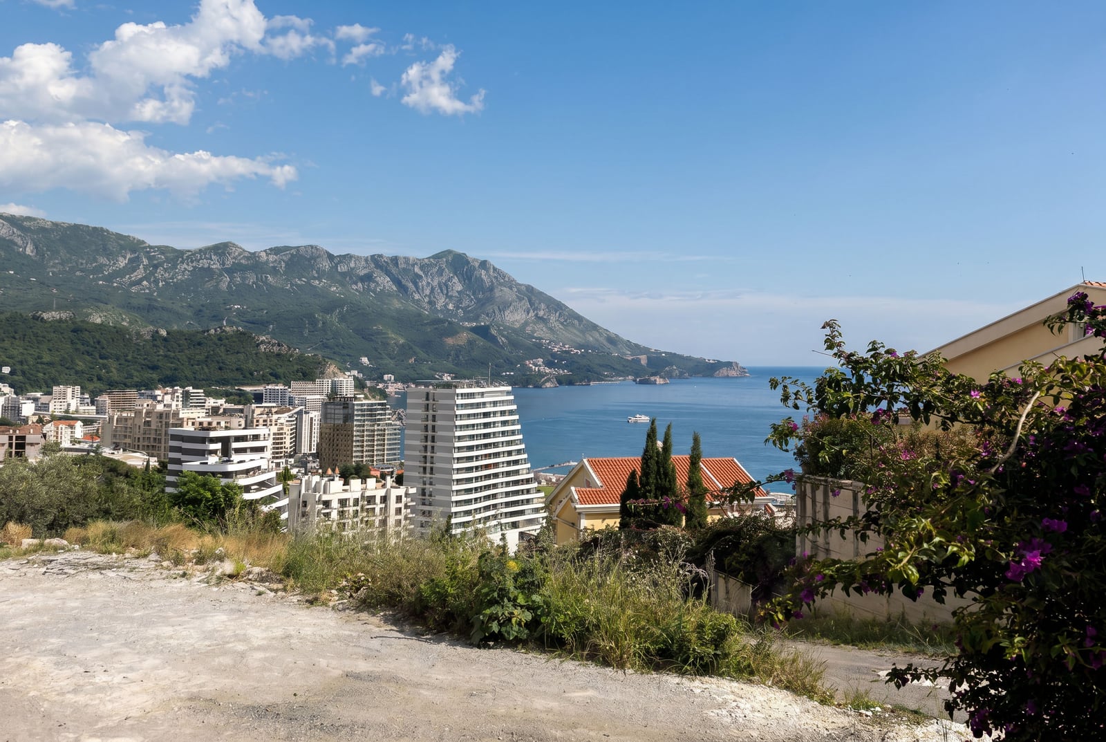 Panoramic view of the sea, mountains, and residential complexes in Bečići, Montenegro, with flowering plants in the foreground.