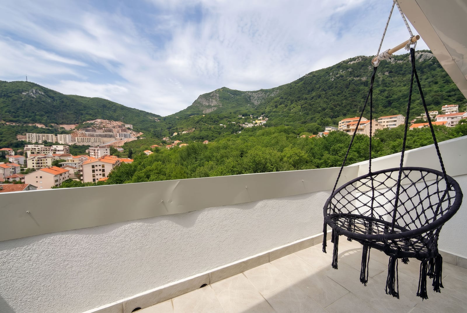 Apartment balcony in Budva, Montenegro, featuring a hanging chair and panoramic views of green mountains and the cityscape.