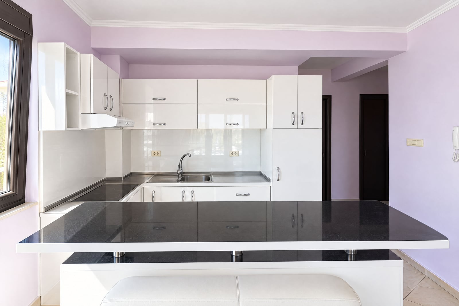 Modern kitchen with white glossy cabinets, a black island countertop, sink, and range hood against light purple walls.