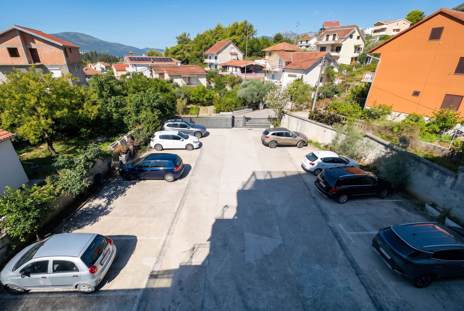 Outdoor parking lot with several cars, surrounded by houses, trees, and distant mountains.