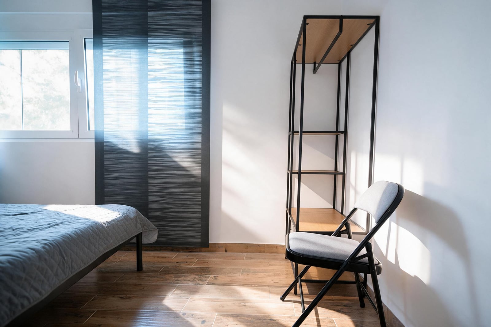 Bedroom featuring a bed with gray cover, black metal shelving unit, and folding chair on wood floor.