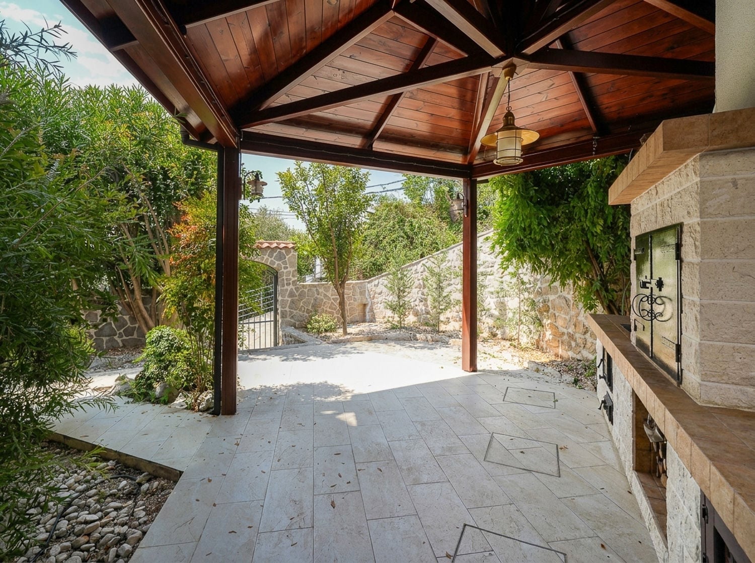 Covered patio with wooden roof, stone BBQ counter, and surrounding garden with stone walls.