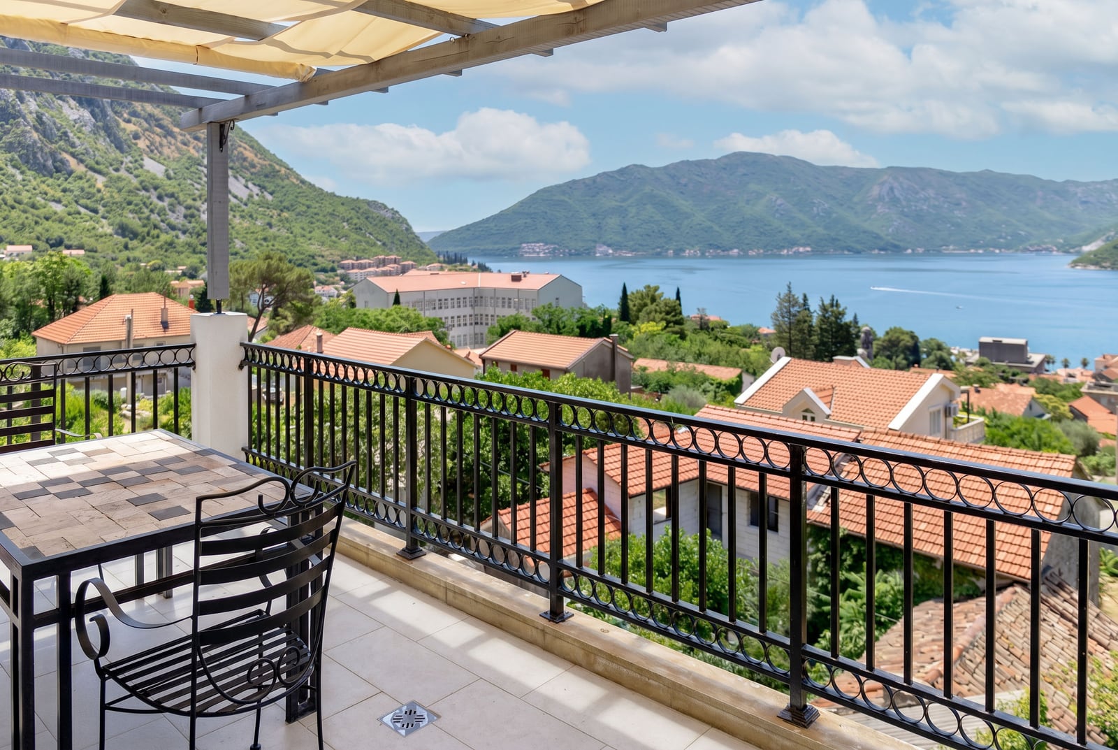 Penthouse terrace in Risan, Montenegro: wrought iron furniture, awning, and panoramic view of the Bay of Kotor and mountains.