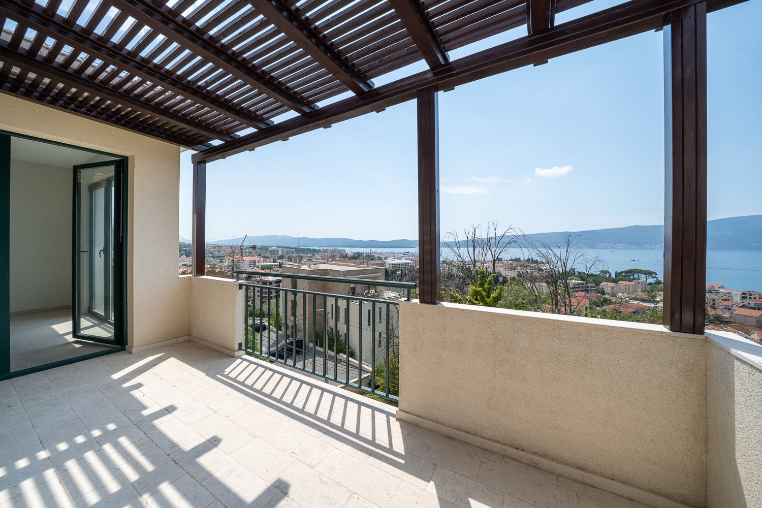 Balcony with wooden pergola and metal railing overlooking a coastal city and sea.