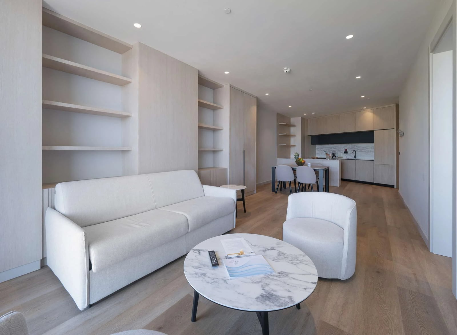 Bright living room with a white sofa, marble table, built-in shelves, and an open kitchen in the background.