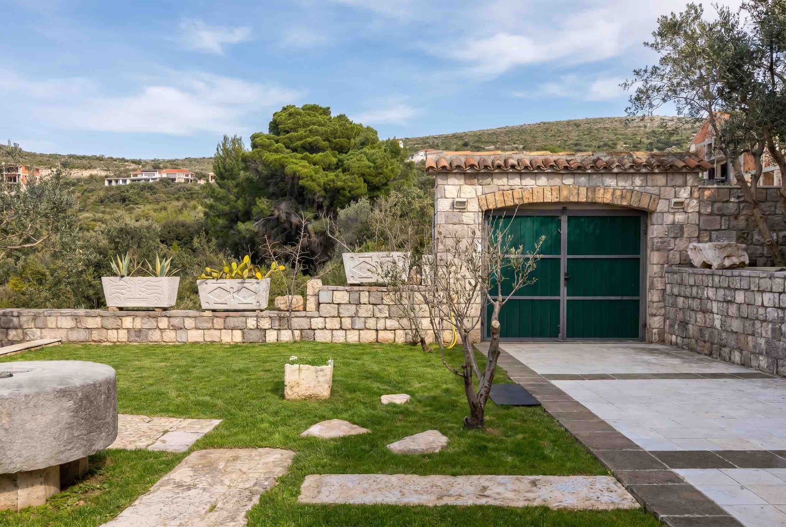 Well-maintained garden with a stone garage and green gates at a house in Luštica, Montenegro, against a backdrop of rolling hills.
