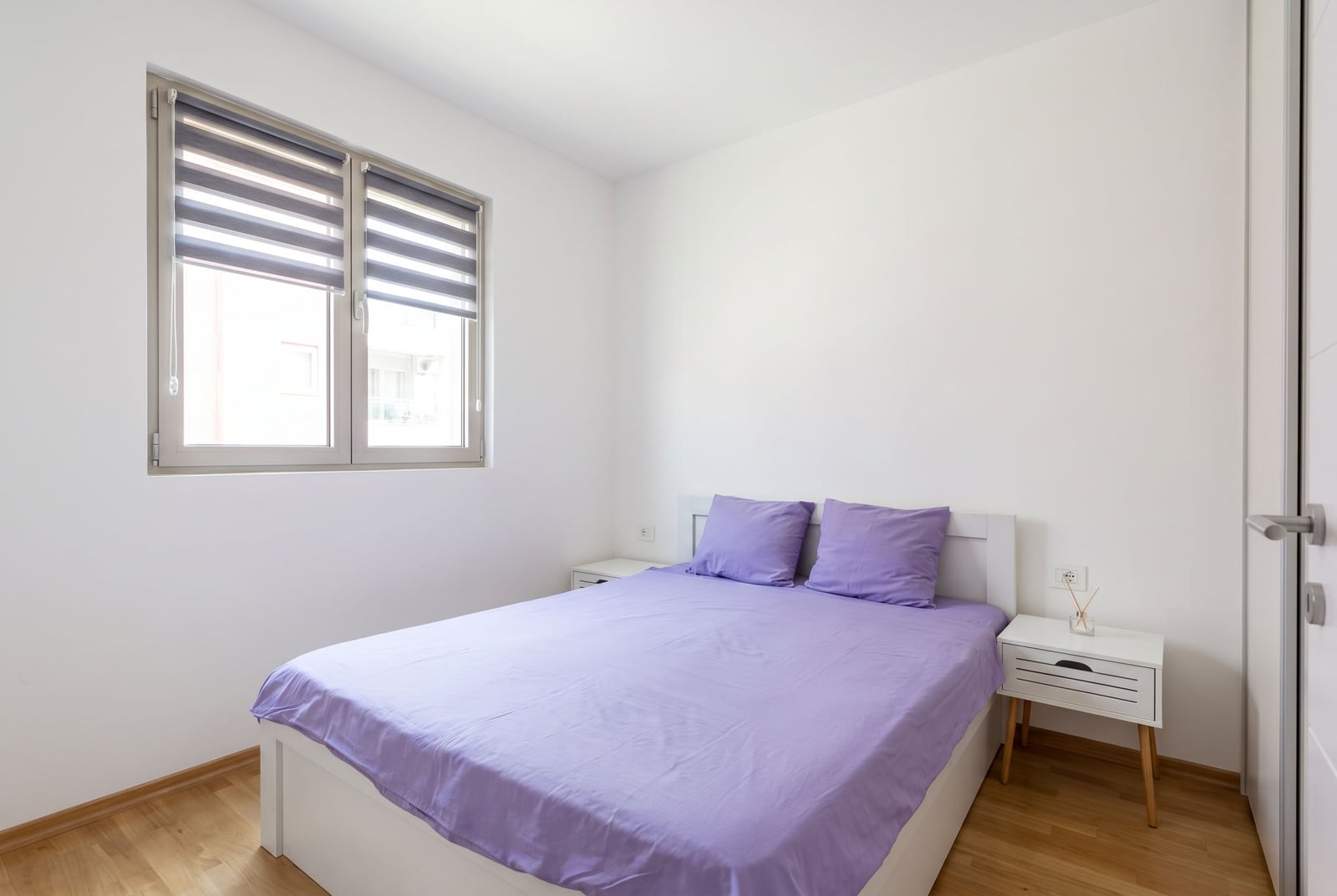 Bright bedroom with a white bed, purple bedding, nightstands, and a window featuring striped roller blinds on light wood floors.