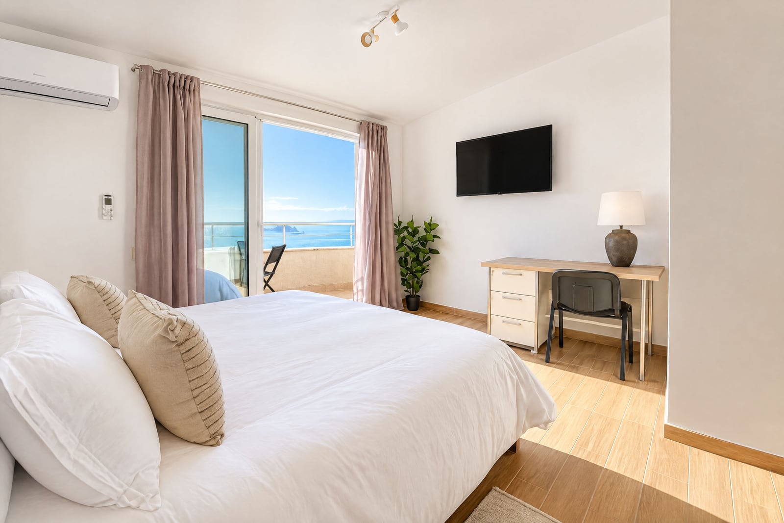 Bright bedroom with a large bed, desk, and TV, featuring a balcony door overlooking the sea.