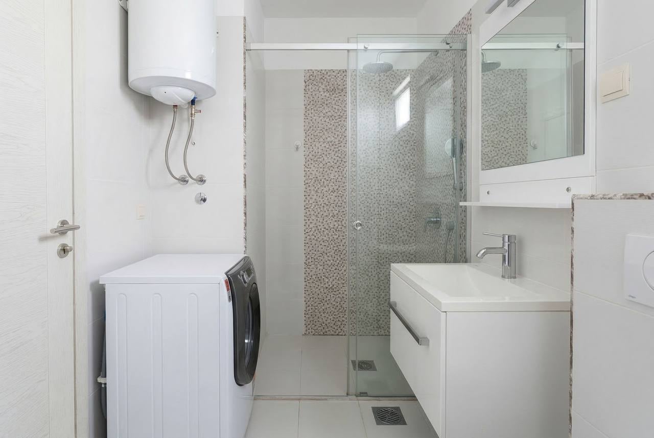 Modern bathroom in an apartment in Bečići, Montenegro: walk-in shower, washing machine, and white furniture.