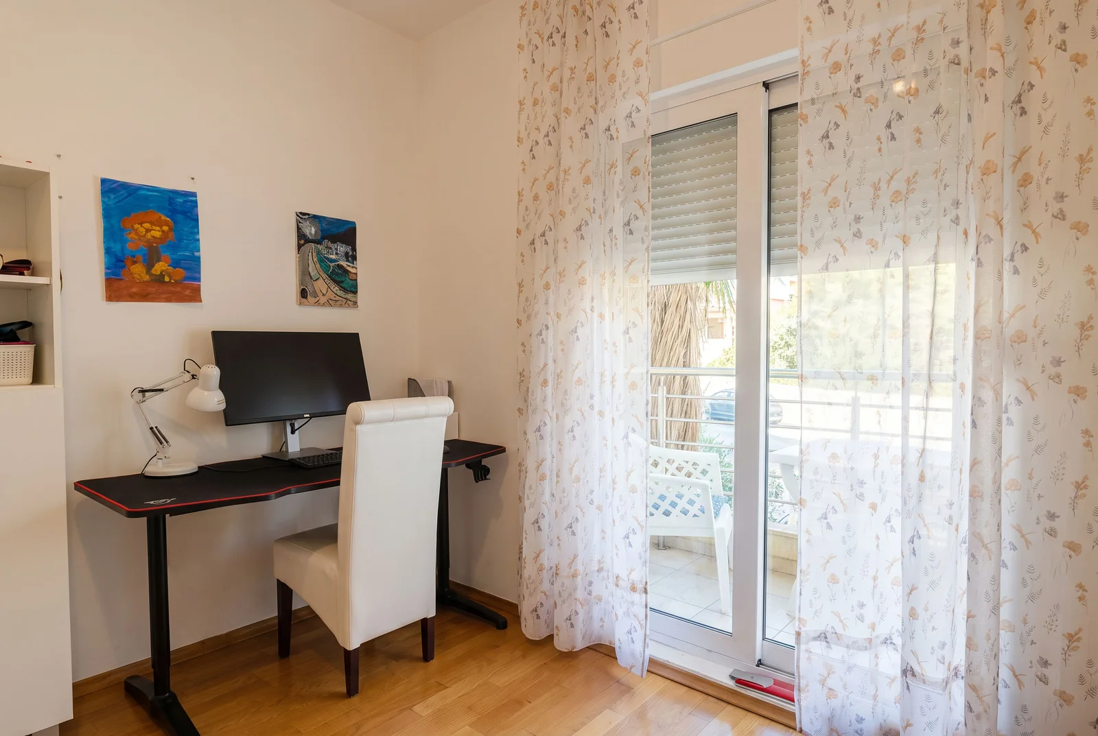 Home office setup with a black desk, white chair, monitor, and floral curtains overlooking a balcony.