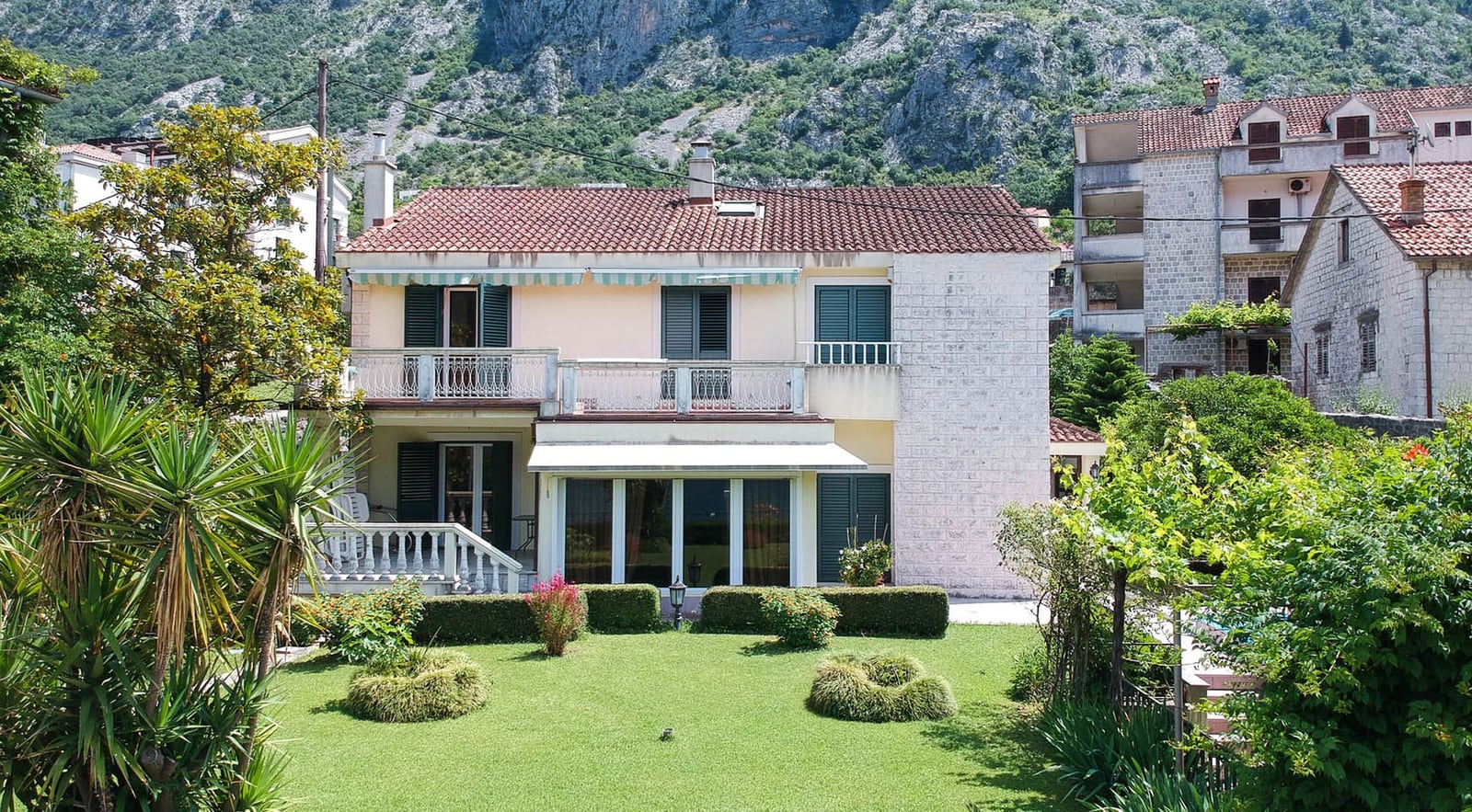 Two-story villa in Dobrota, Montenegro: a house with a terrace, panoramic windows, a green garden, and mountain views.