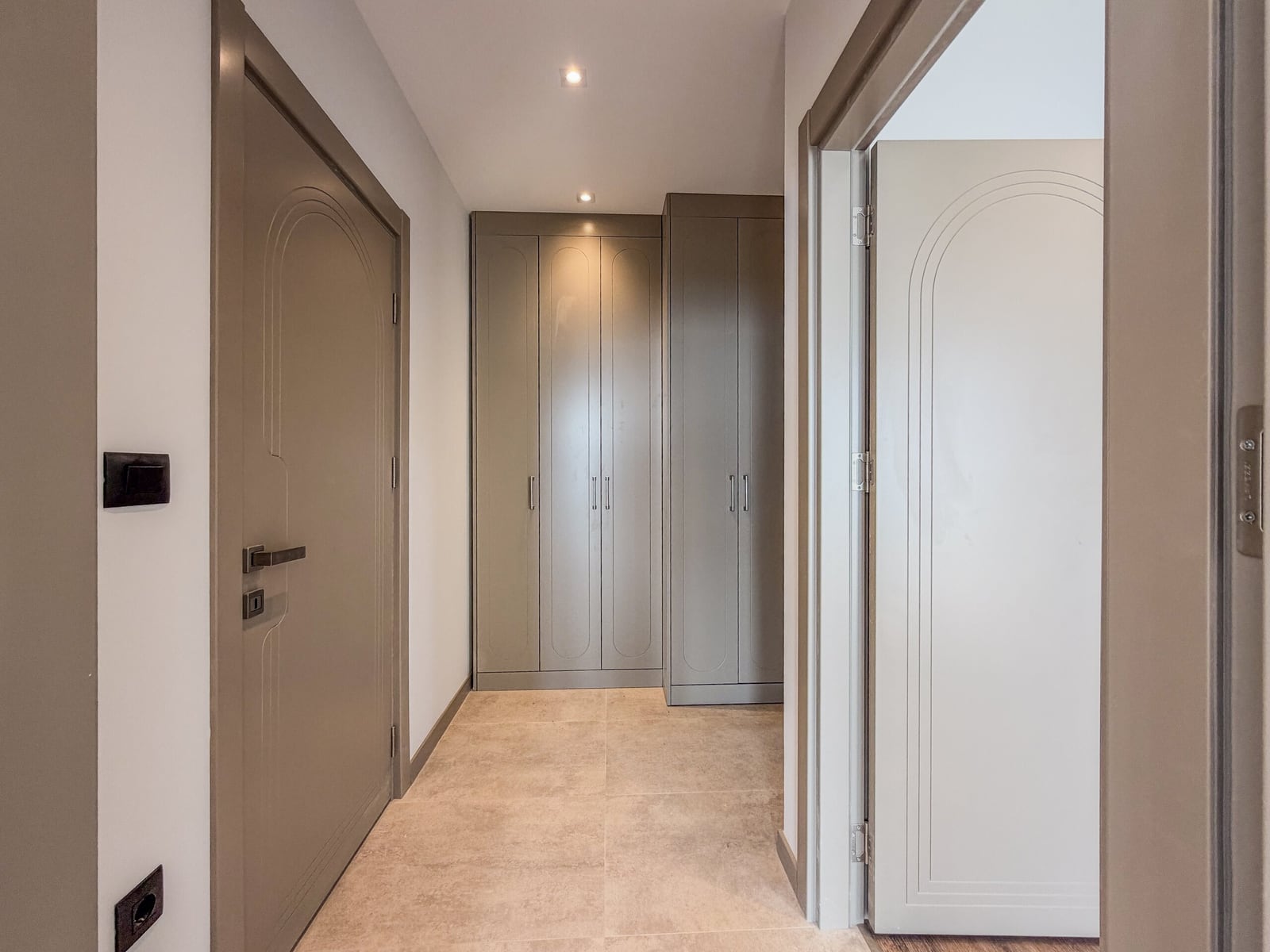 Hallway with grey built-in wardrobes, beige floor tiles, and paneled doors under recessed lighting.