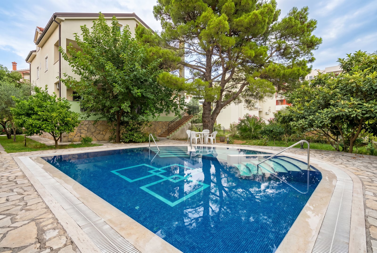 Outdoor swimming pool with a lounge area at a villa in Budva, Montenegro, surrounded by a green garden and tall pine trees.