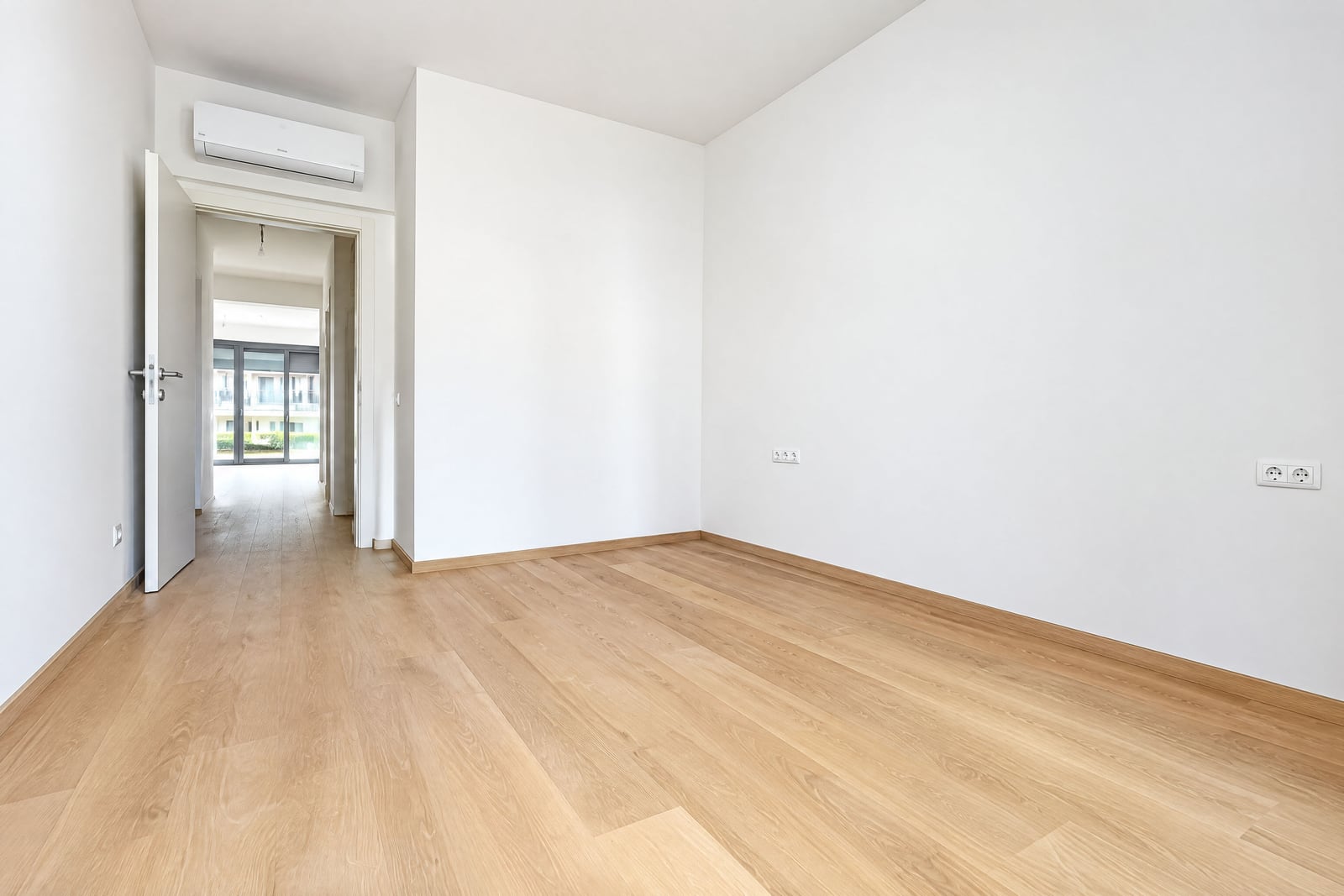 Empty room with wooden flooring and AC unit, open door leads to a hallway.