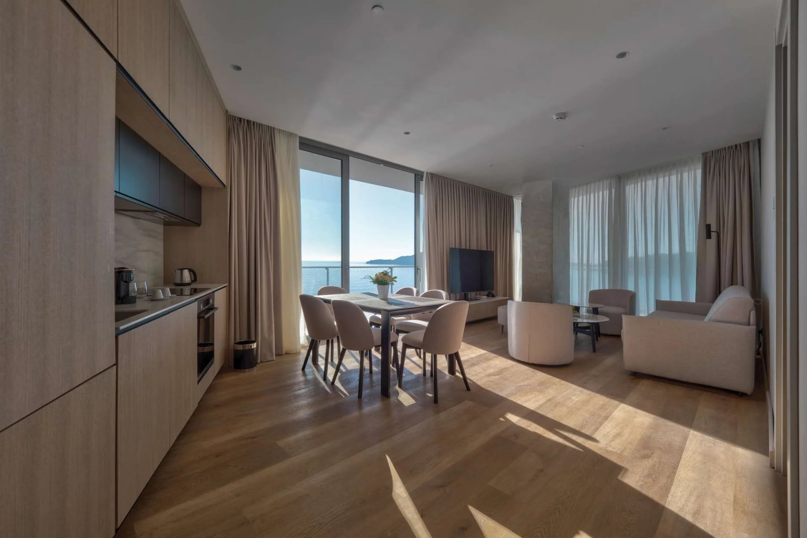 Open-plan living room with kitchenette, dining table, and sofa facing a large window with sea view.