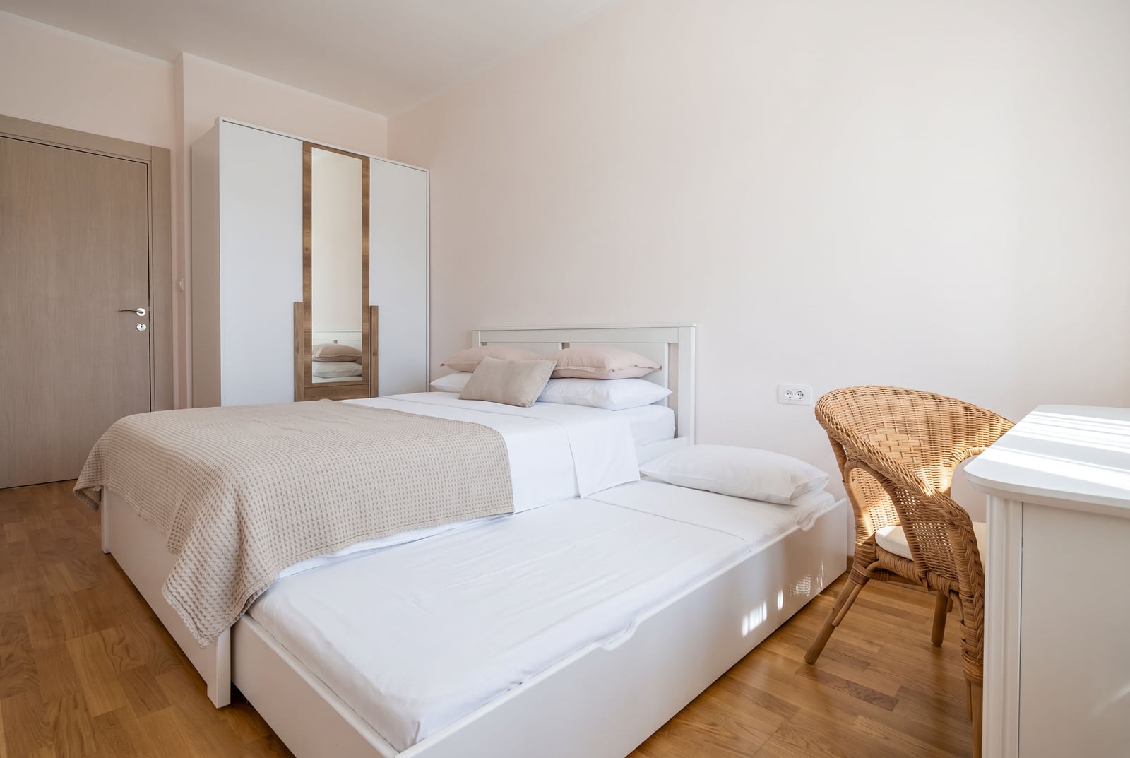 Bright bedroom in a Budva apartment, Montenegro: double bed with a pull-out trundle, mirrored wardrobe, and a wicker chair.