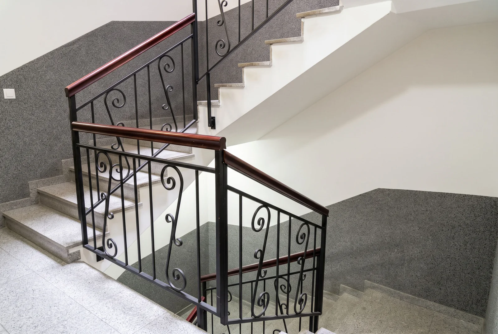Stone staircase featuring ornate black wrought iron railings with a dark wood handrail against a textured grey wall.