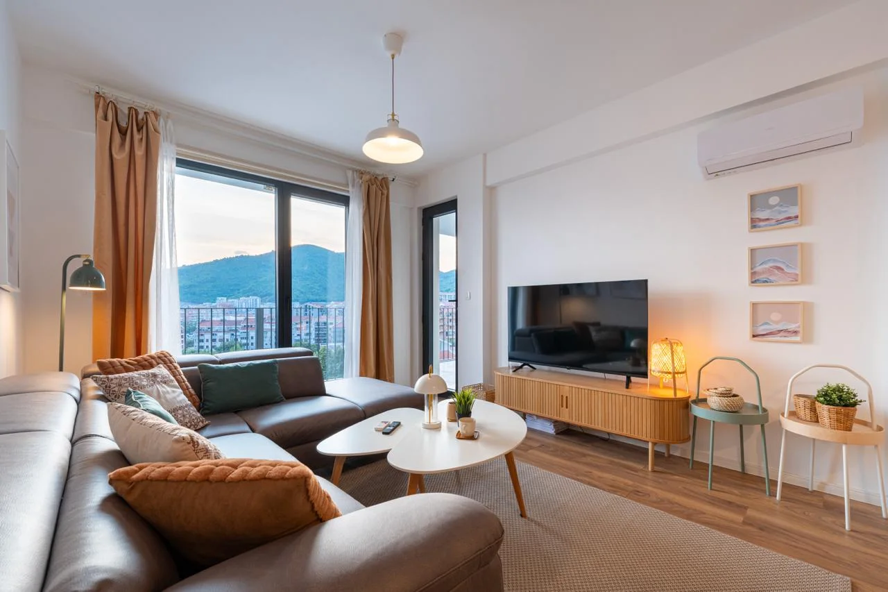 Bright living room with a grey sofa, wooden TV stand, and large window offering a mountain view.