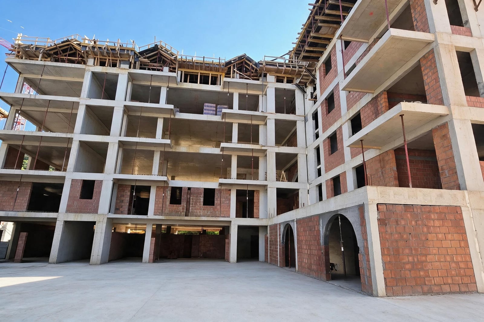 Unfinished multi-story building construction showing concrete frame, brick walls, and wooden roof formwork under a clear blue sky.