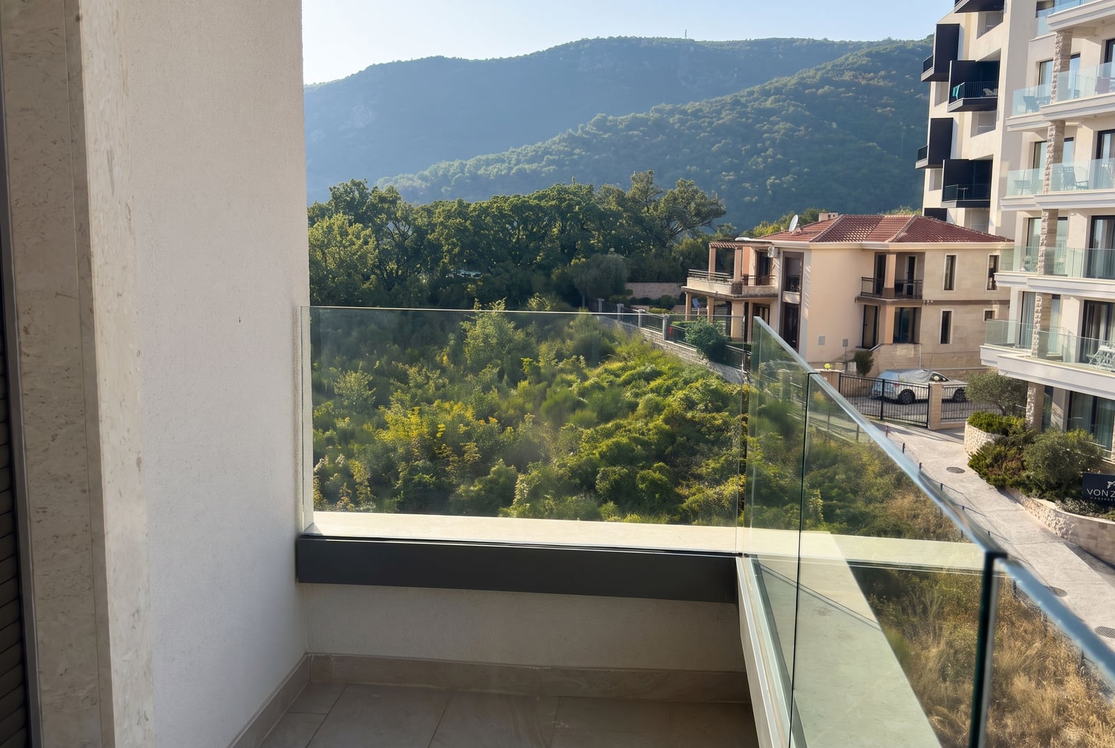 Balcony with glass railing overlooking green vegetation and mountains. Part of an adjacent building is visible.