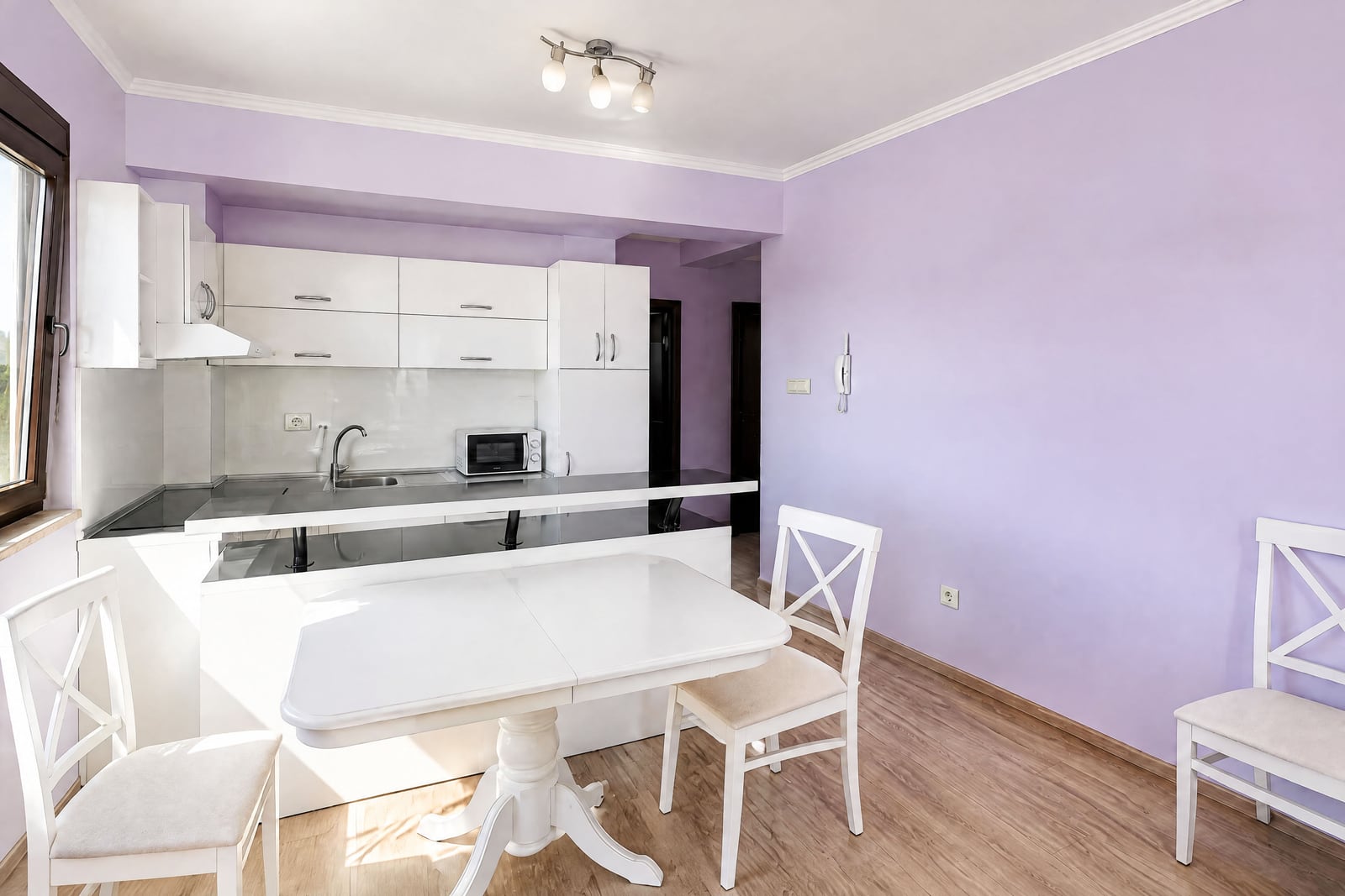 Kitchen with white cabinets, black countertop, sink, microwave, and dining table against lavender walls.
