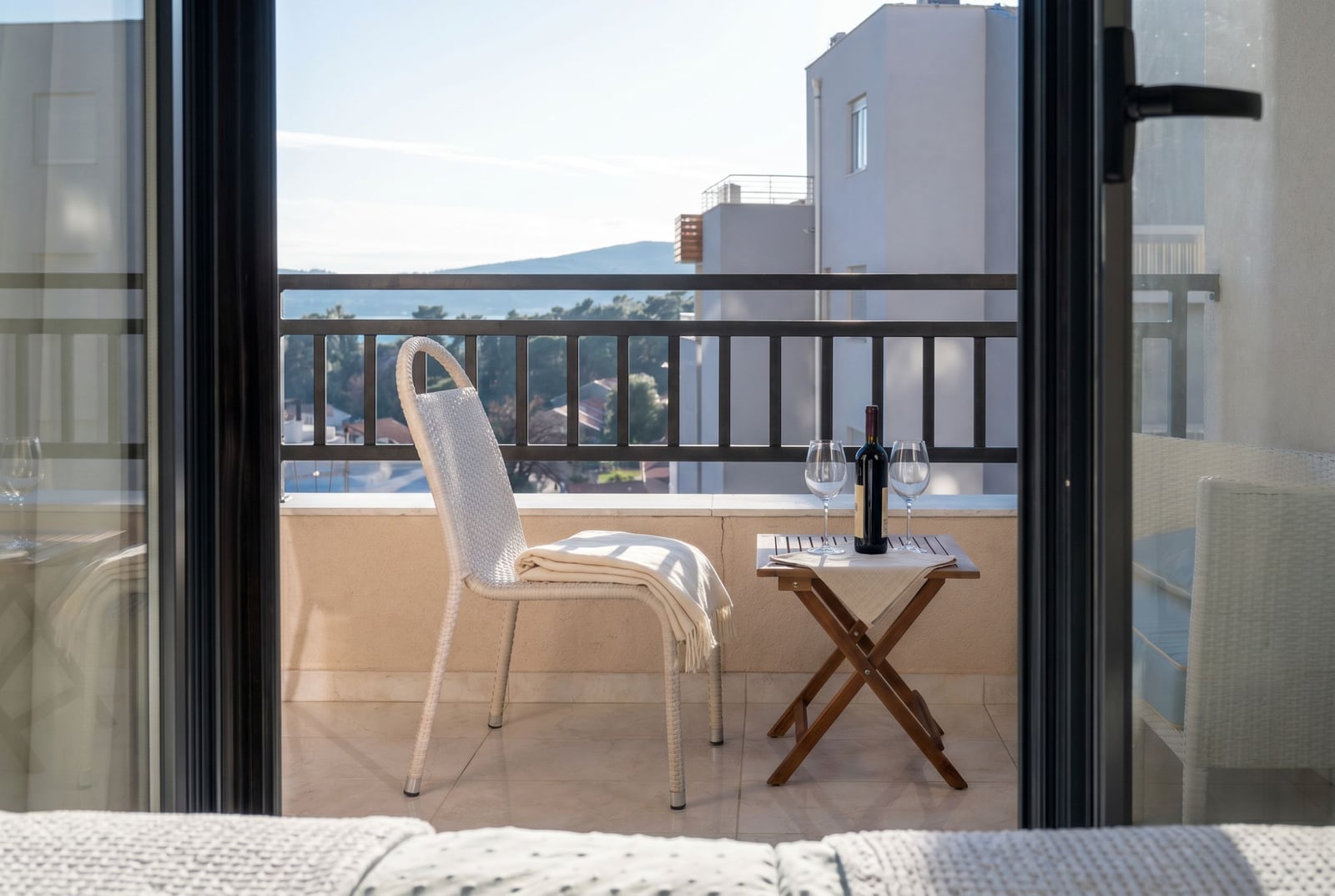 Apartment balcony in Tivat, Montenegro: wicker furniture, a table with wine, and a panoramic view of the sea and mountains.