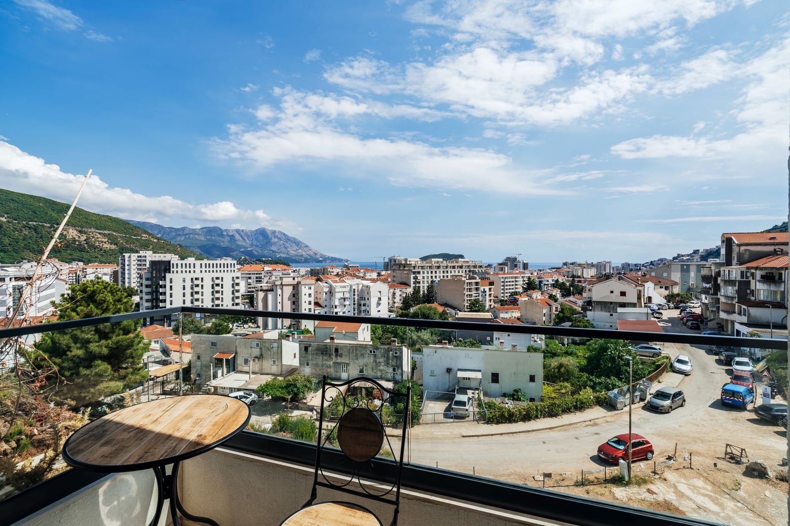 View from the apartment balcony in Budva, Montenegro: panoramic view of the city, mountains, and sea, with a table and chair on the terrace.
