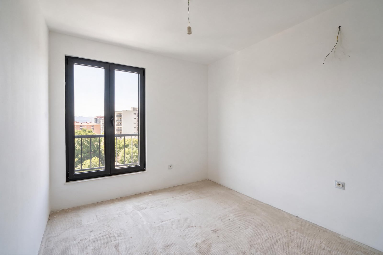 Empty room with white walls, black window frame, unfinished floor, exposed wires and outlets visible.