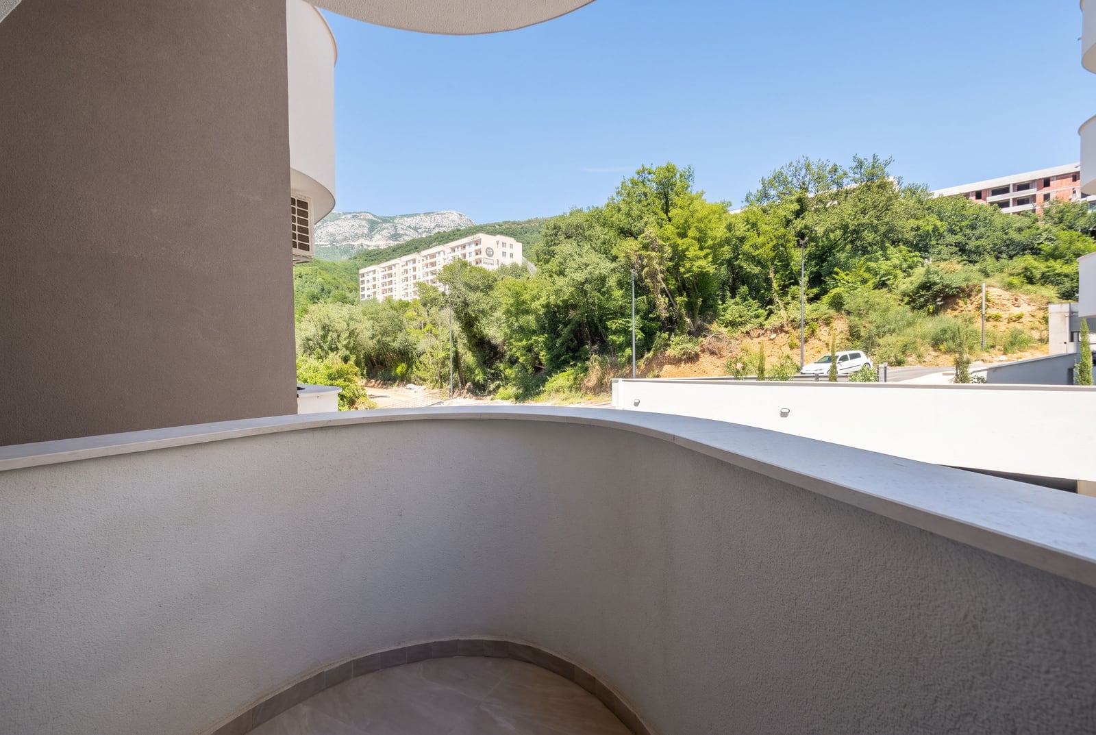 Curved balcony with a grey parapet overlooking green hills, trees, and other buildings under a clear blue sky.