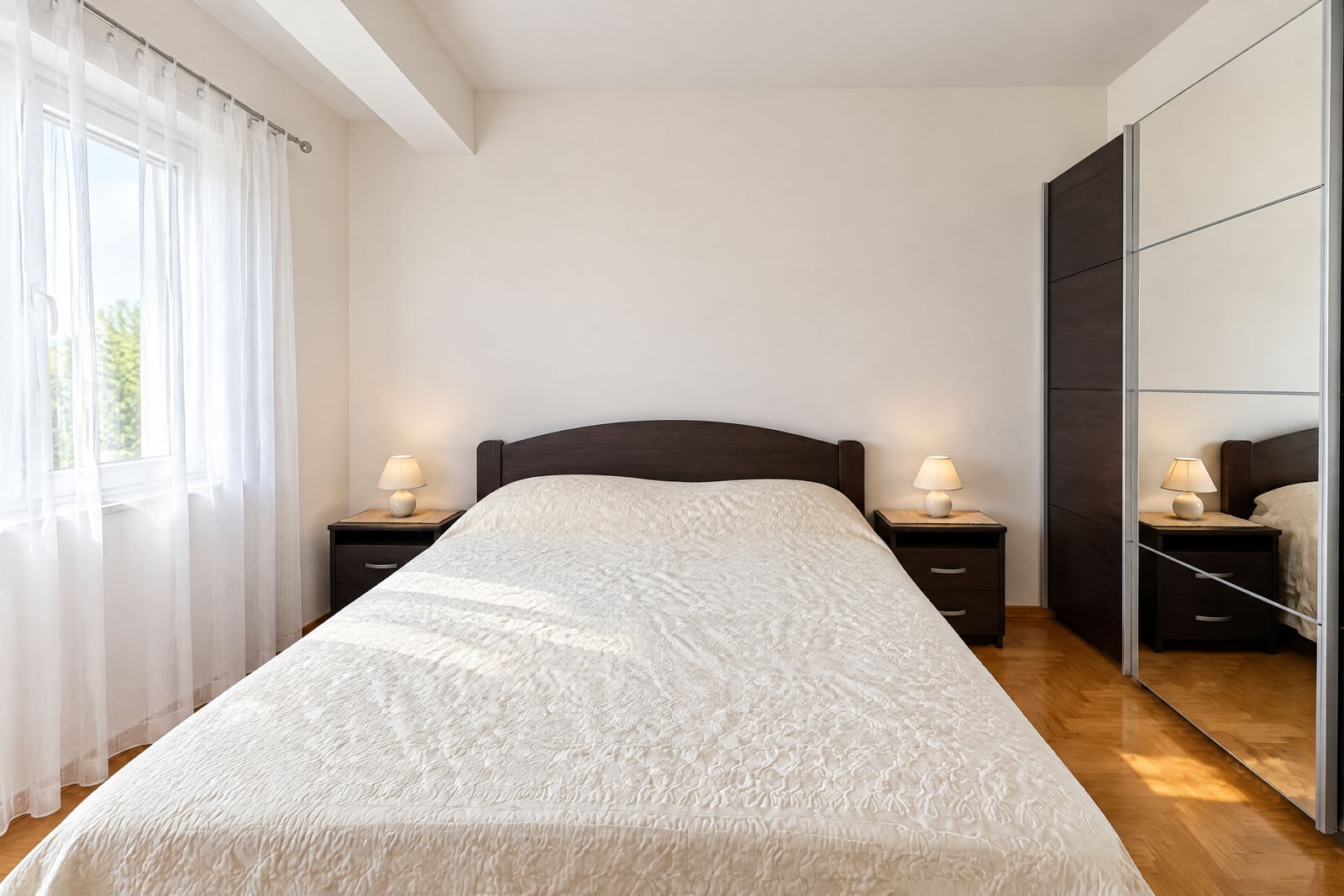 Bedroom featuring a double bed with white cover, dark wood nightstands, lamps, and a large mirrored wardrobe sliding door.