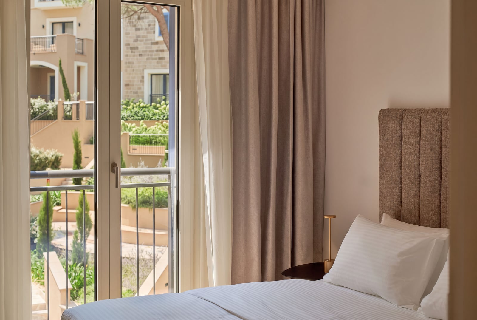 Bed with white linens and upholstered headboard next to a glass door opening to a balcony.