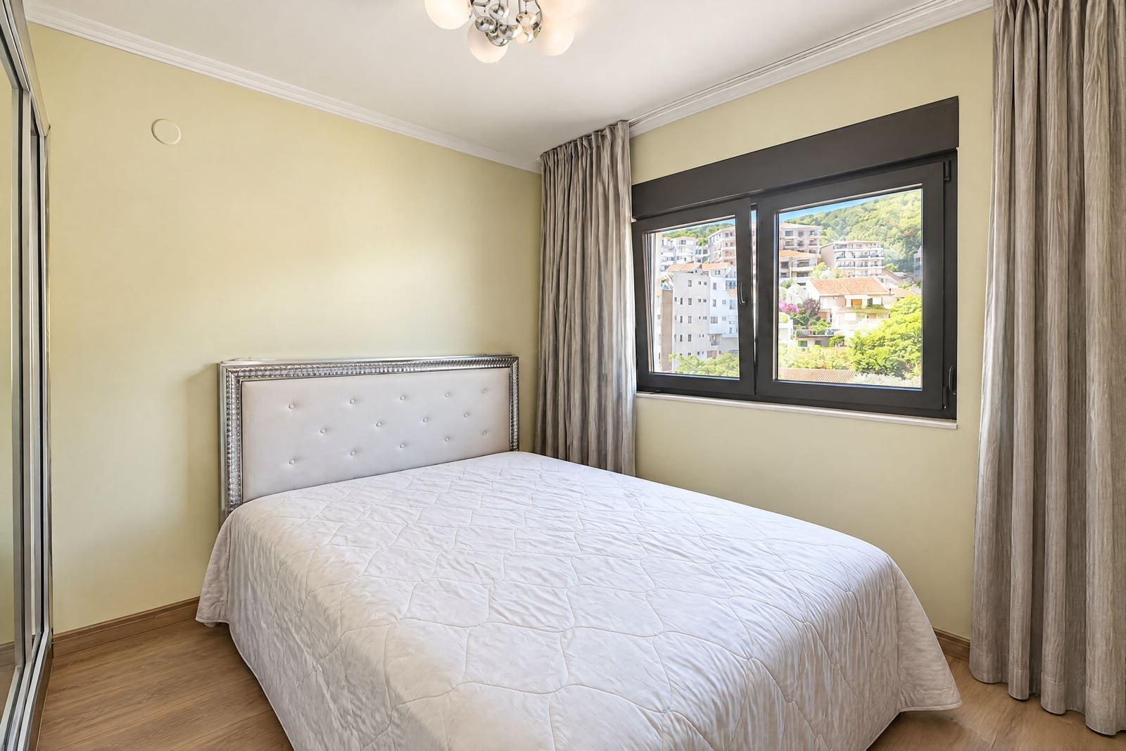 Bedroom featuring a white bed with an upholstered headboard, beige walls, wood flooring, and a window.