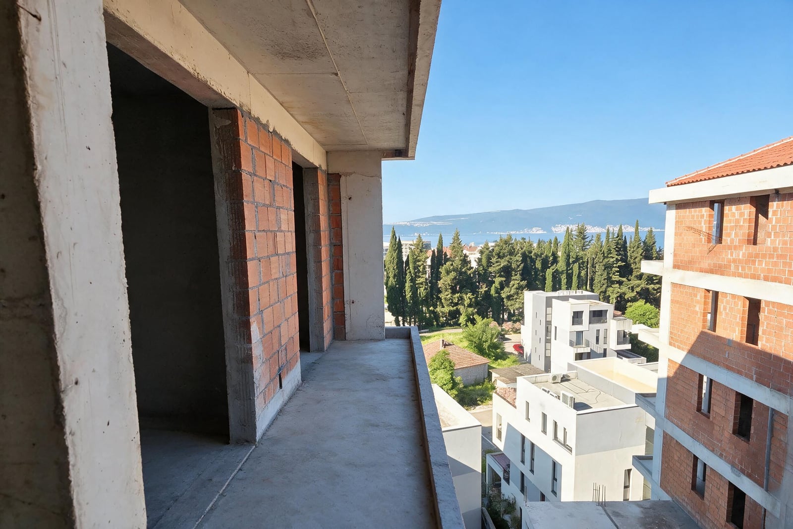 Concrete balcony in an unfinished red brick building overlooking the sea and trees.