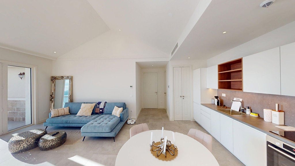 Modern living room with kitchen in a Lustica Bay apartment, Montenegro: blue sofa, white furniture, and terrace access.
