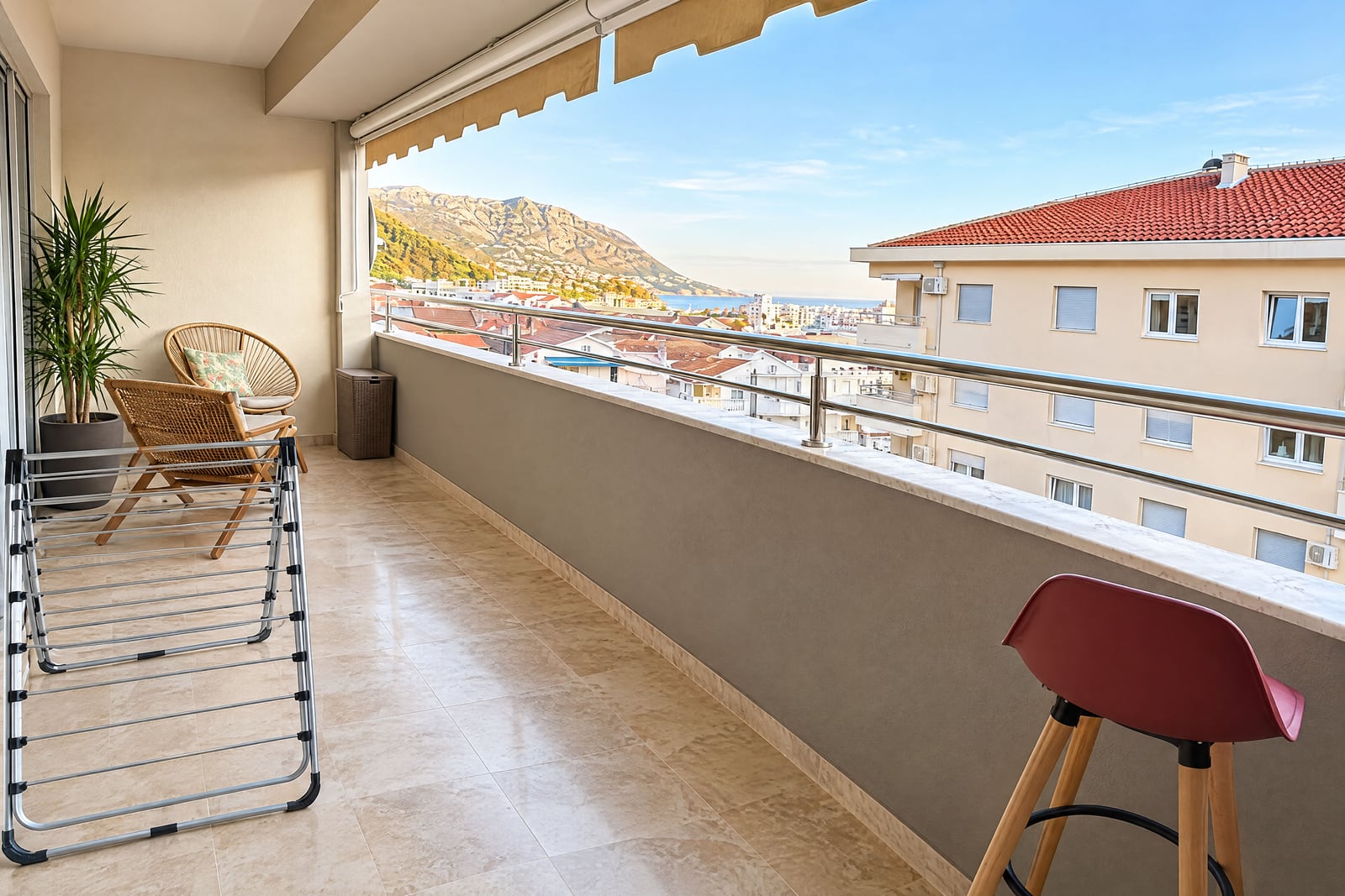 Wide tiled balcony with wicker chairs, a drying rack, and a scenic view of the city and mountains.