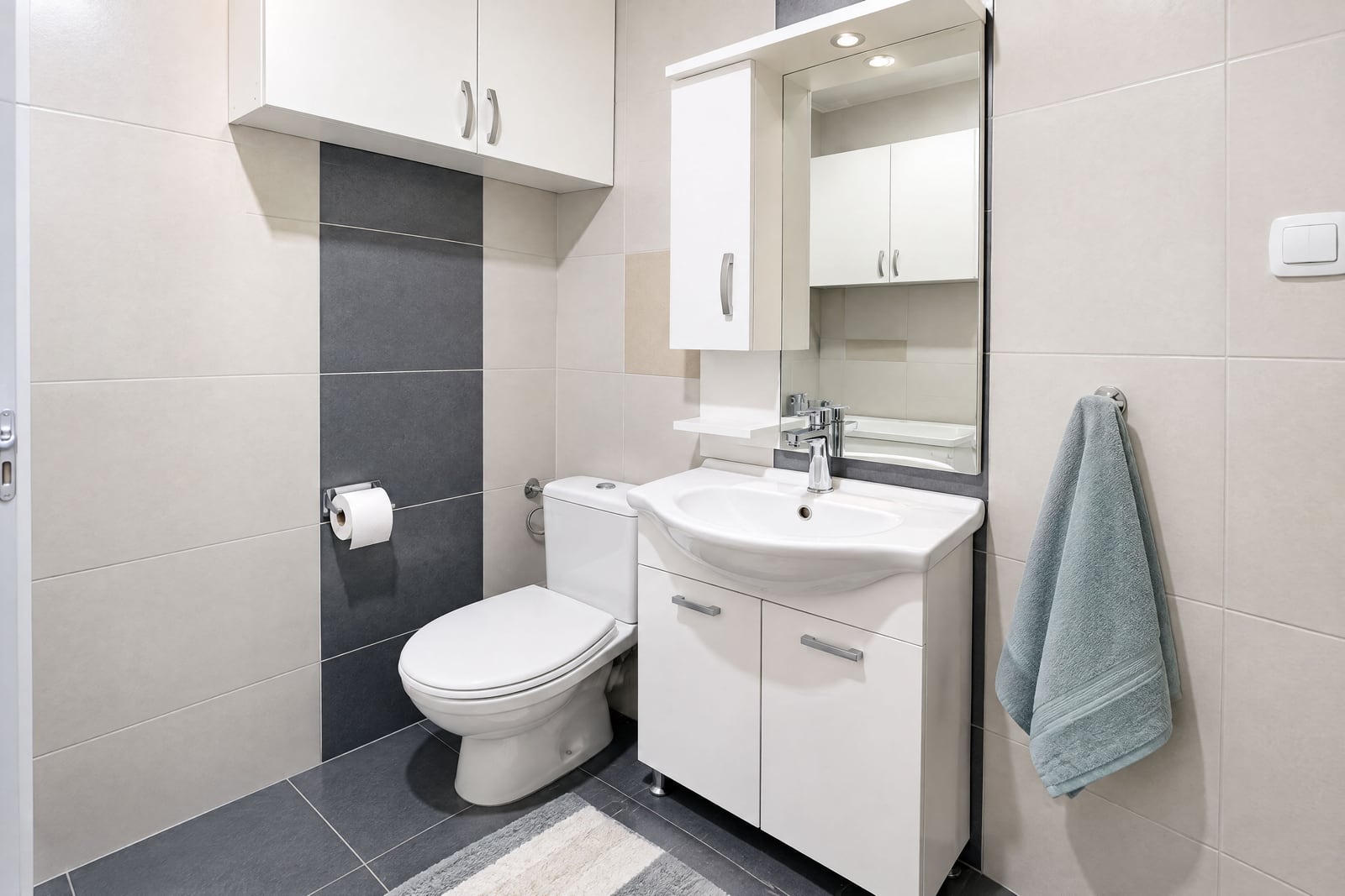 Modern bathroom featuring a white toilet, vanity sink with mirror, wall cabinets, and two-tone grey and beige wall tiles.