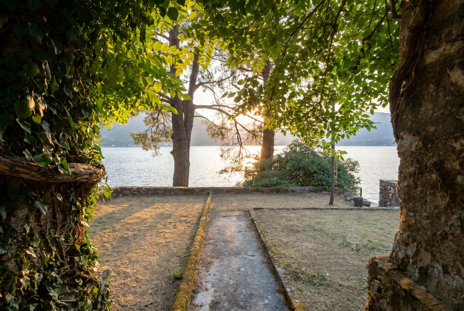 View of the sea and mountains from a property in Dobrota, Montenegro: green grounds, trees, and a stone path leading to the shore.