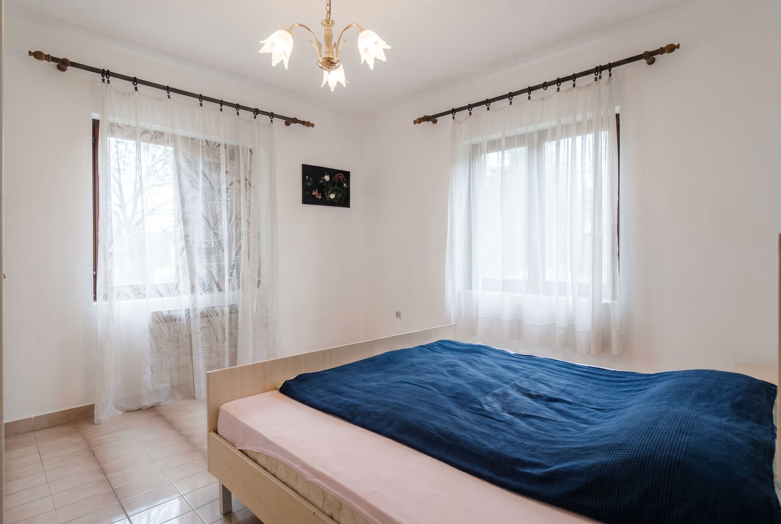 Bright bedroom with a double bed in a house for sale, Lastva, Montenegro: tiled floor and two windows with sheer curtains.