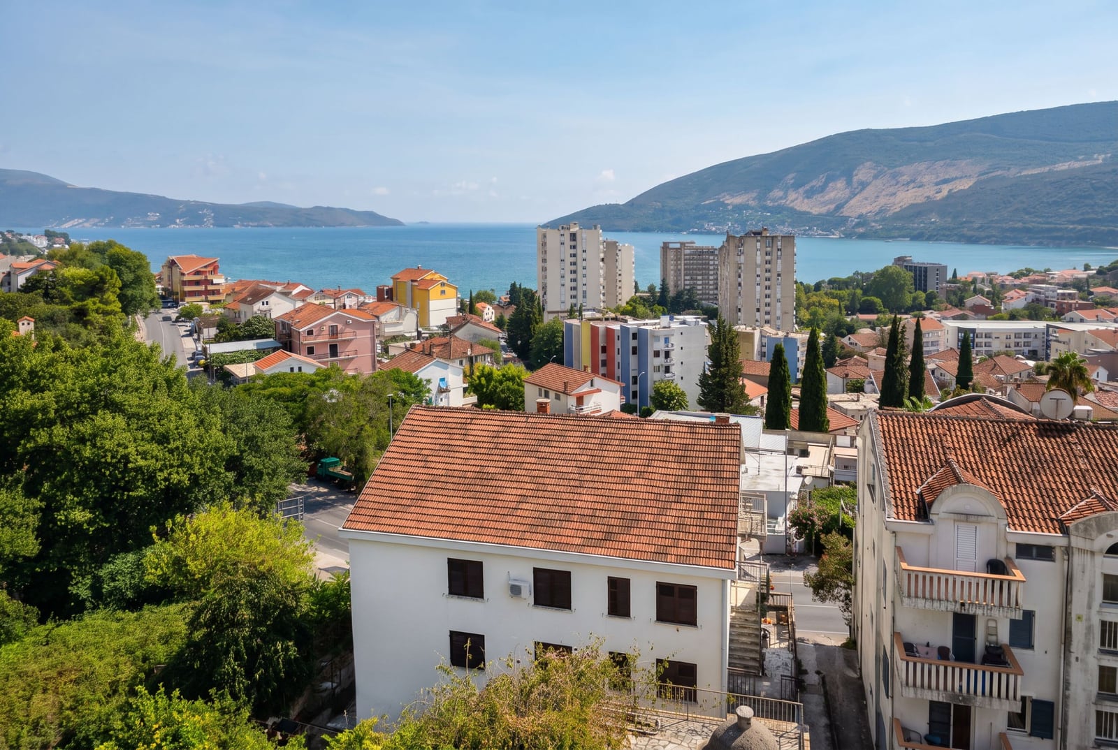 Panoramic view of city rooftops, sea, and mountains from an elevated exterior vantage point.