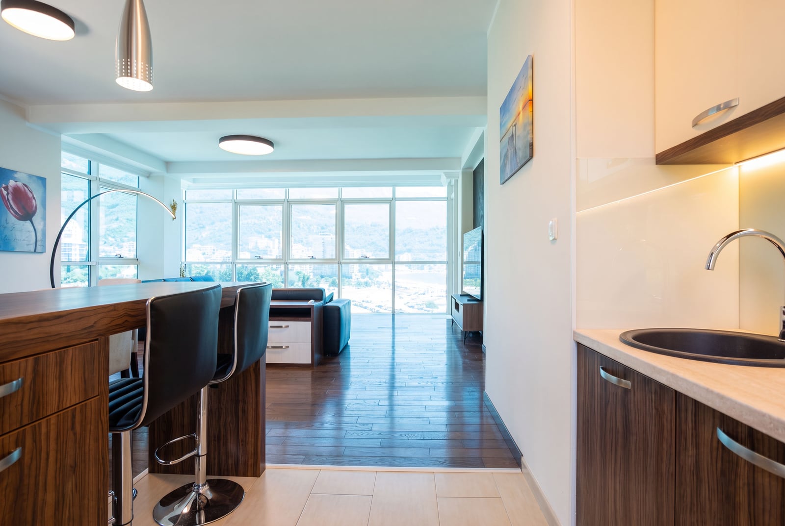 Kitchen area with a wooden bar counter and black sink, adjacent to a living room with a sofa and large windows.