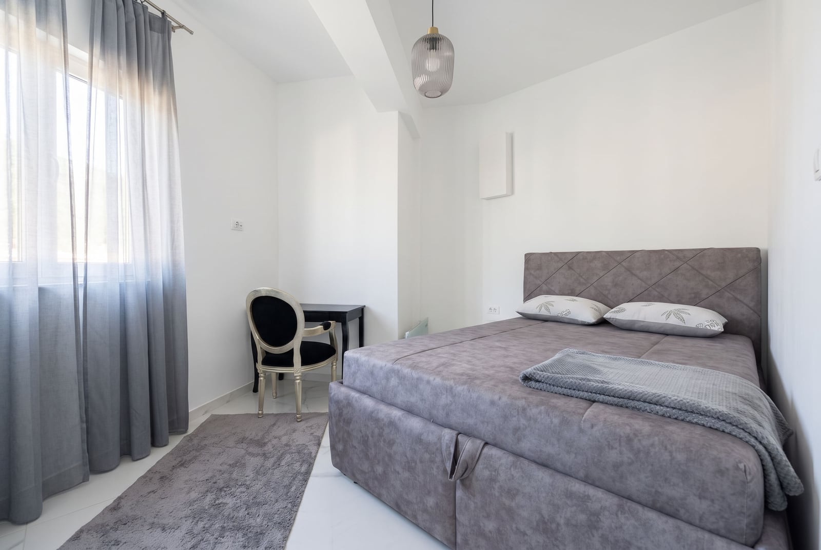 Bright bedroom featuring a grey upholstered bed, an ornate chair by the window, and a pendant light.