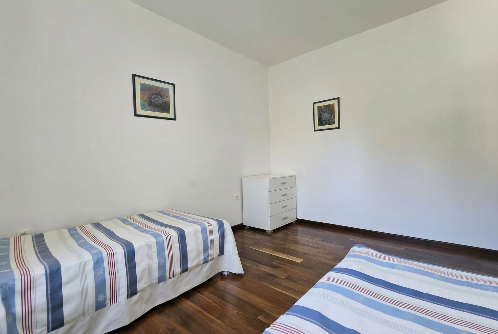 Bright bedroom with two beds and a white dresser in an apartment in Tivat, Montenegro, with dark wood flooring.