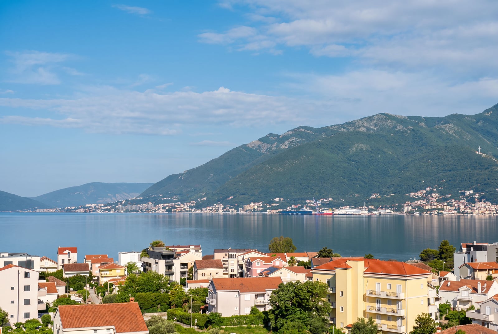 Panoramic view of a coastal town with red roofs, sea, and green mountains under a blue sky.