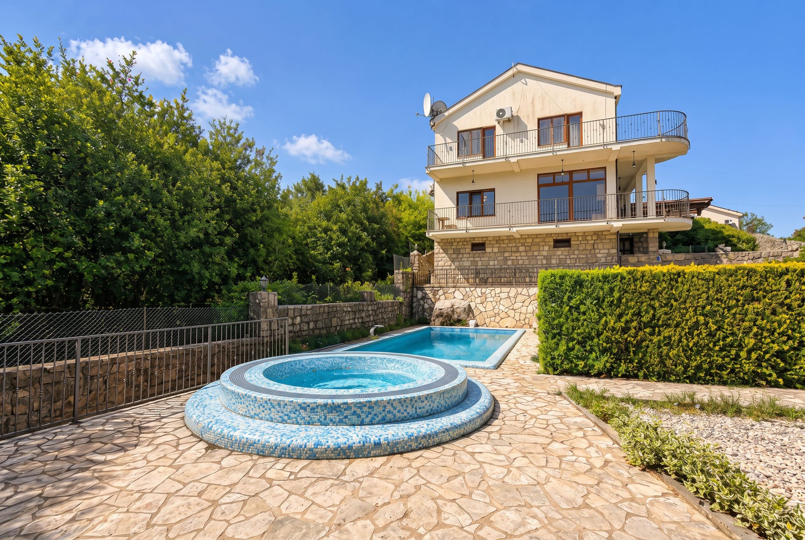Round mosaic jacuzzi and rectangular swimming pool on a stone patio in front of a two-story house.