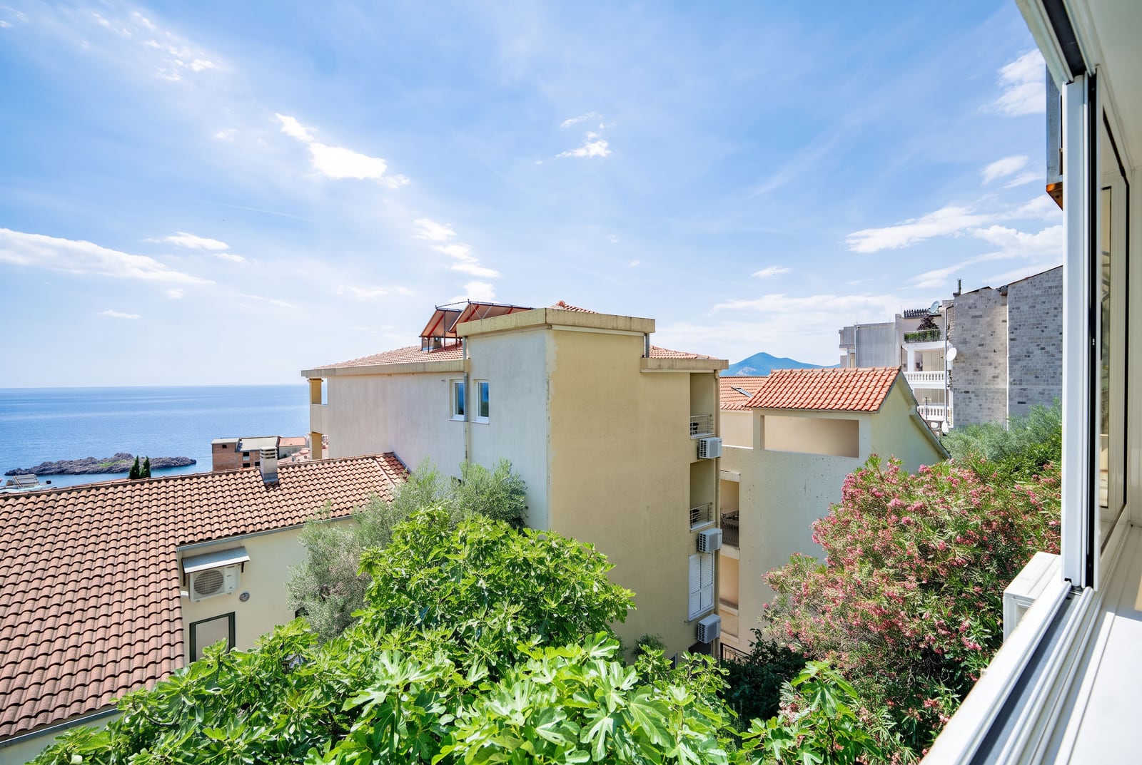 View from the balcony of an apartment in Sveti Stefan, Montenegro: panorama of the sea, tiled roofs, and a blooming garden.