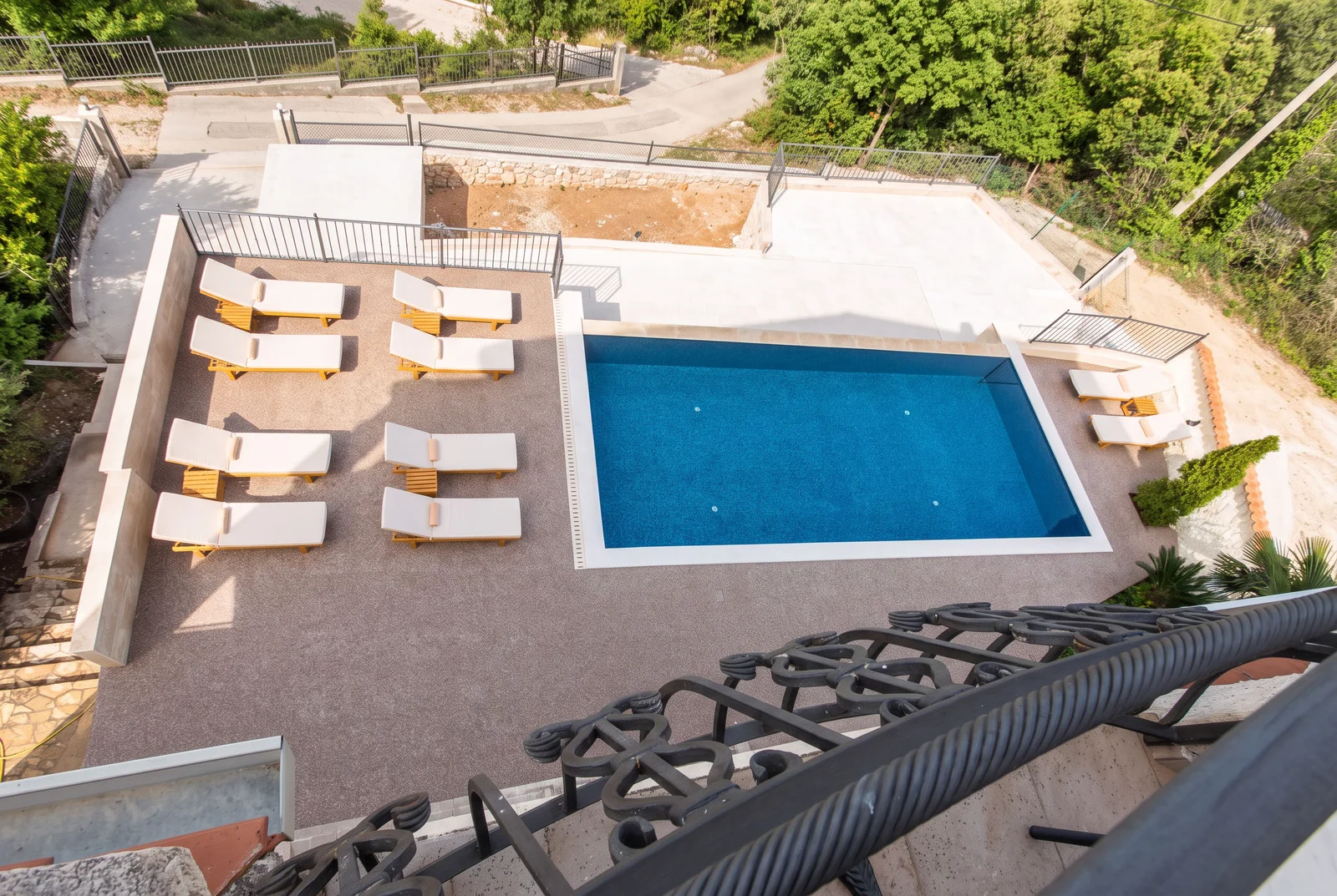 High-angle view of a blue swimming pool with white sun loungers on a patio surrounded by greenery and fencing.