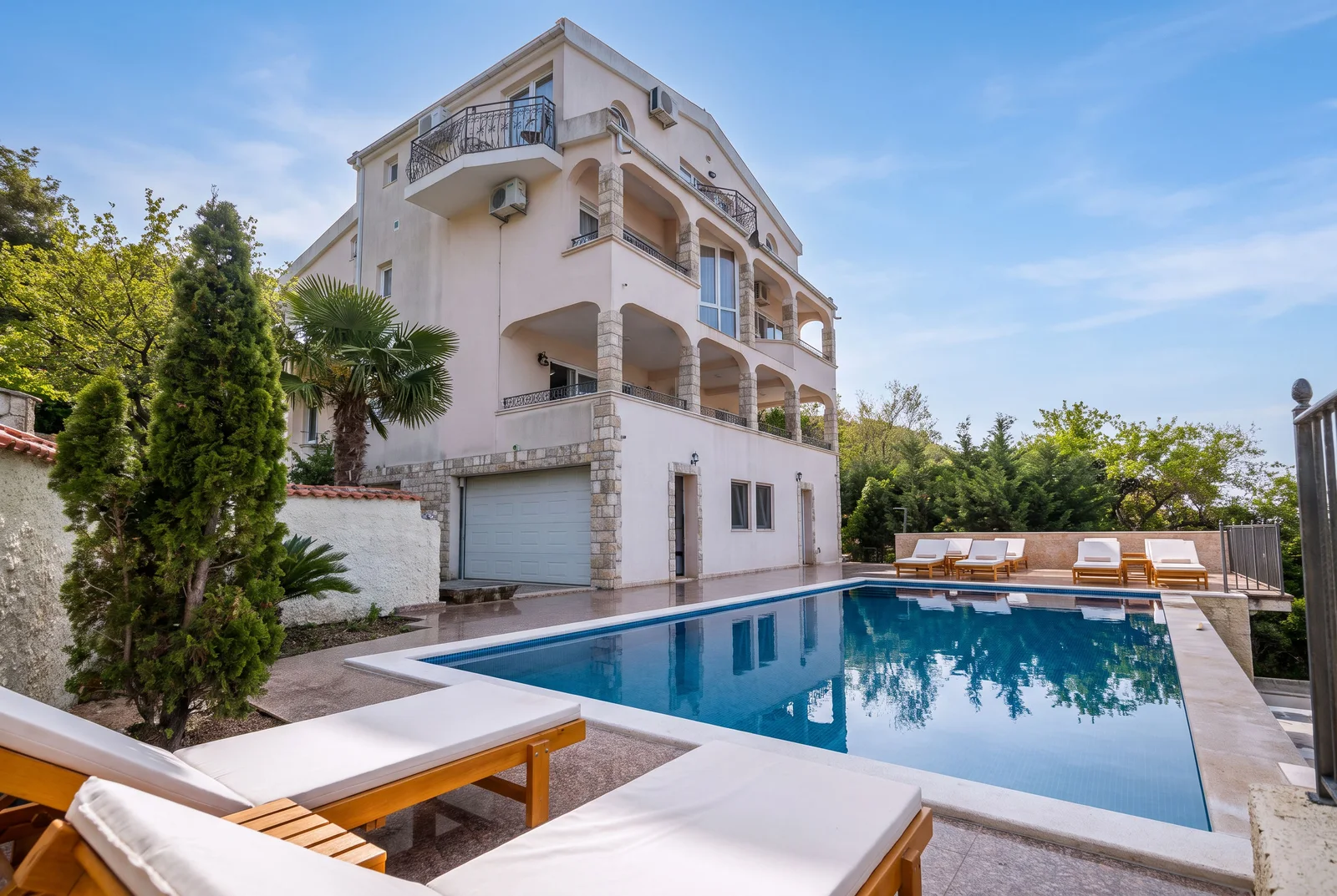 Private swimming pool with wooden loungers and a multi-story house surrounded by trees.