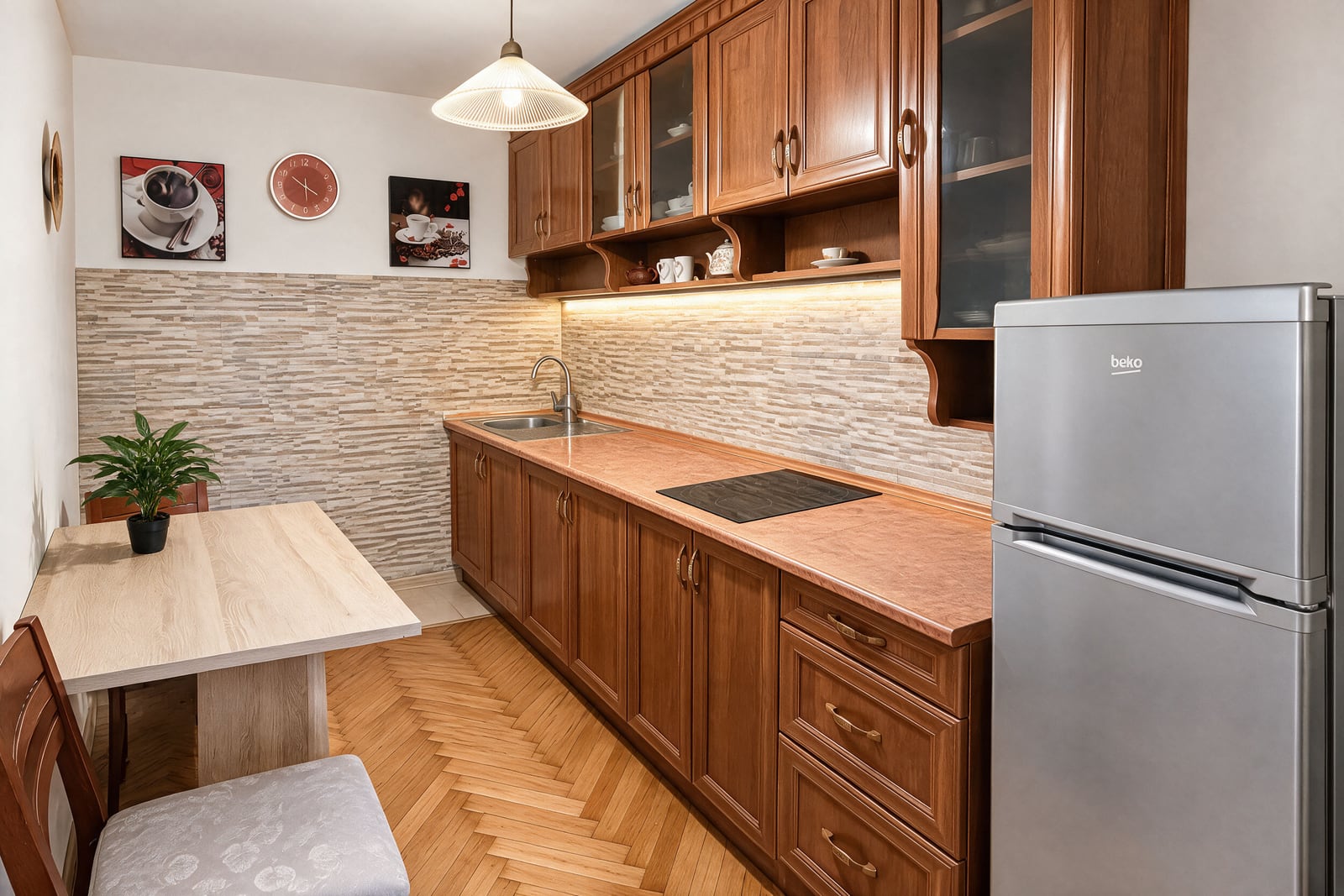 Kitchen featuring wooden cabinets, a silver Beko fridge, sink, induction cooktop, and a small dining table.