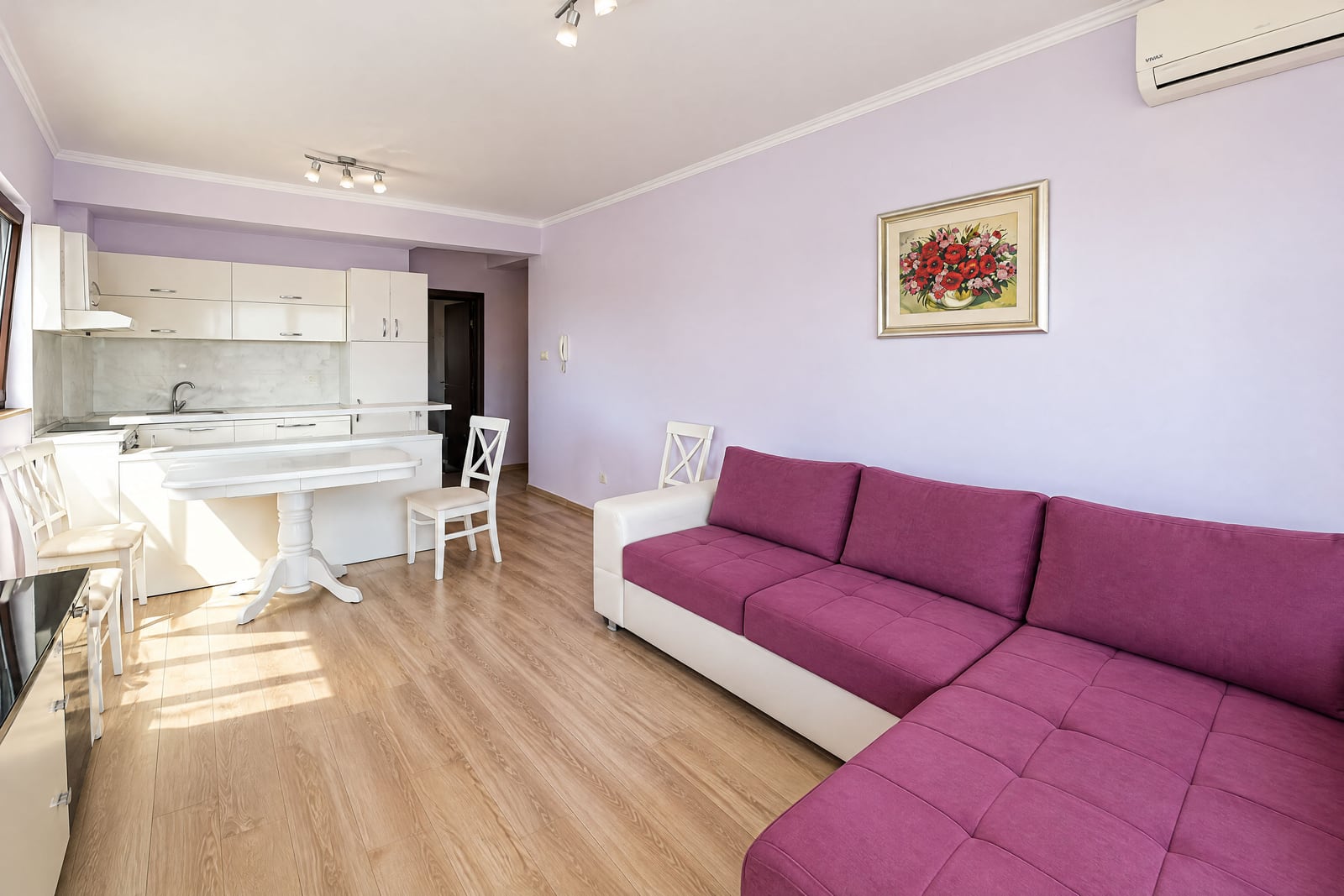 Living room with a purple sofa and white kitchen area on light laminate flooring.